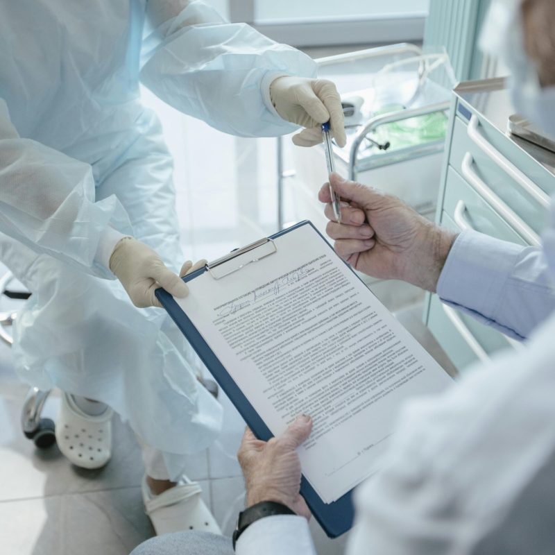 Healthcare workers in protective gowns exchanging a pen over a clipboard in a hospital room
