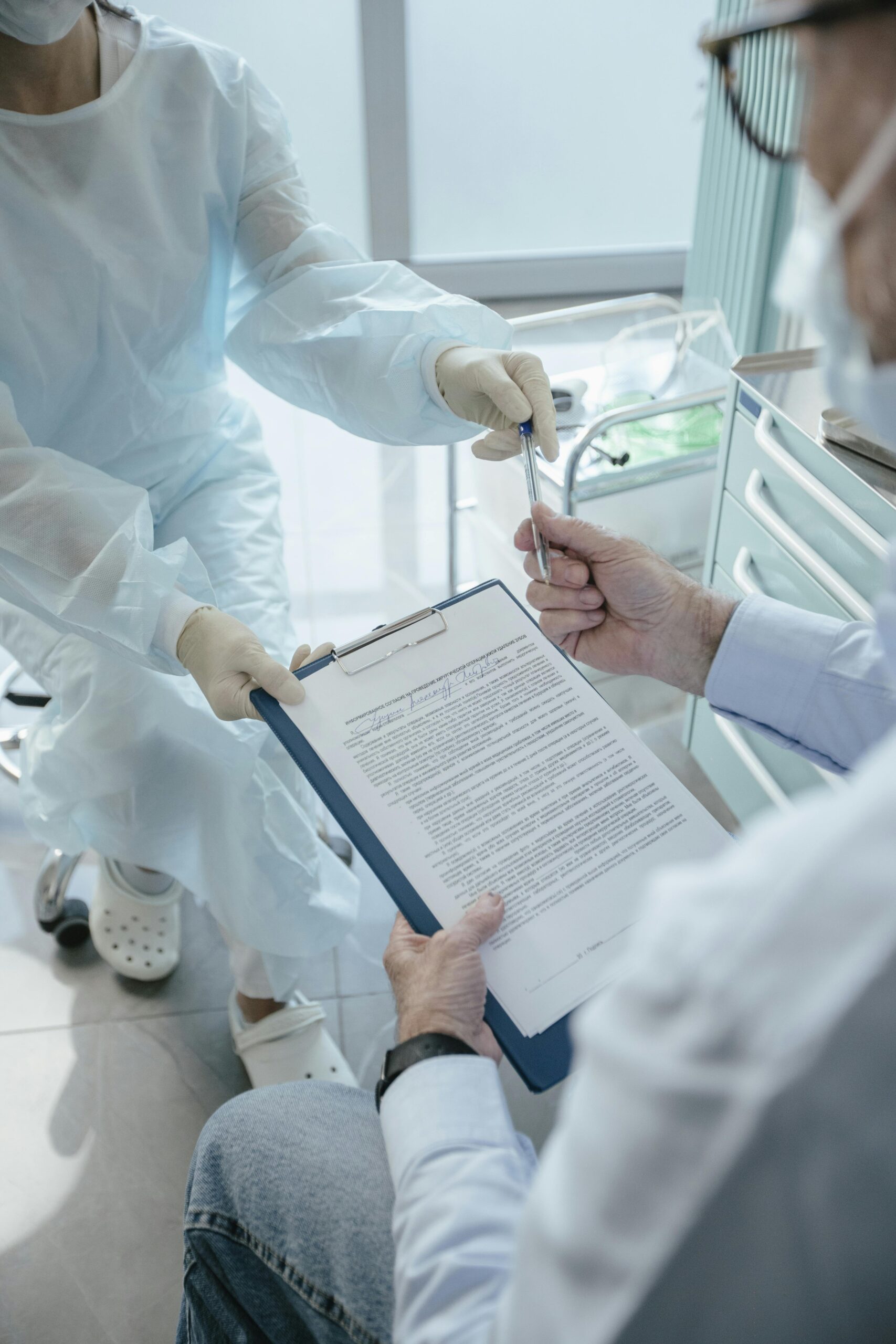 Healthcare workers in protective gowns exchanging a pen over a clipboard in a hospital room